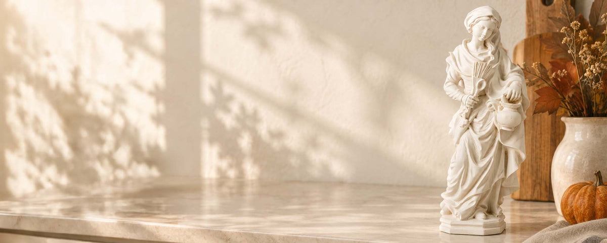 A white classical-style figurine of a robed figure holding a child stands on a light stone surface against a warm beige wall with soft leaf shadows. A ceramic vase with dried stems and a small pumpkin sit beside it.